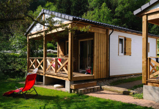 Modern glamping cabin with wooden porch and red lounge chair, surrounded by greenery in a holiday park.