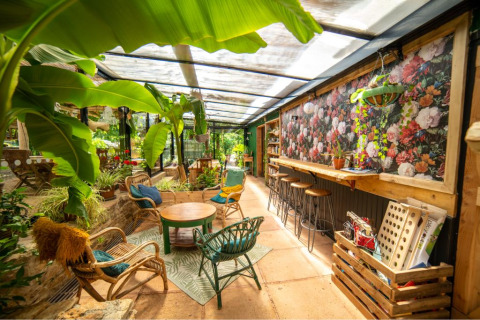 Soleada terraza acristalada con plantas, muebles de mimbre y mural floral en Camping Les 3 Cantons, Occitania.