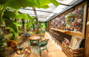 Bright, colorful sunroom filled with greenery, rattan furniture, and a floral wall at Camping Les 3 Cantons.