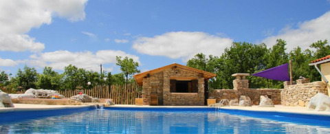 Piscina con edificio in pietra e alberi sullo sfondo al Camping Les 3 Cantons, Occitanie, Francia.
