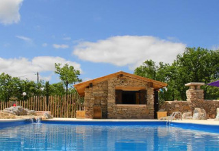 Piscina con edificio in pietra e alberi sullo sfondo al Camping Les 3 Cantons, Occitanie, Francia.