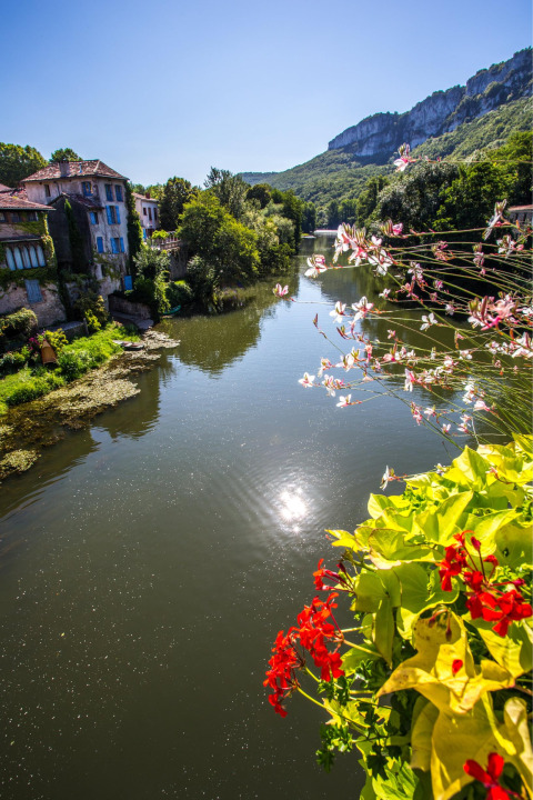 Vista sul fiume con fiori, case tradizionali e colline al Camping Les 3 Cantons, Occitanie, Francia.