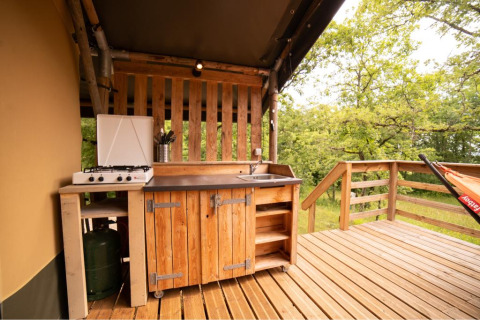 Buitenkeuken op het terras van een safaritent met gasvuur, houten kasten, spoelbak en zicht op het bos.