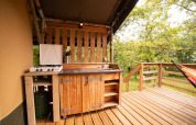 Buitenkeuken op een safaritent terras met gasfornuis, houten kastjes, gootsteen en bosuitzicht.