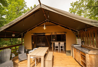 Safari tent with wooden furniture, outdoor kitchen, and seating area on a covered deck among trees.