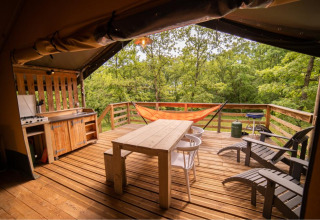 Tienda safari con terraza de madera, hamaca, cocina exterior, mesa, sillas y vista al bosque verde.
