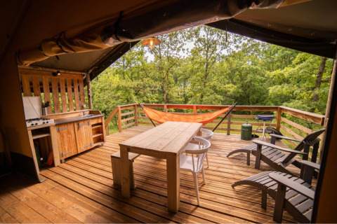 Tenda safari con terrazza in legno, amaca, cucina all'aperto, tavolo, sedie e vista sul bosco.