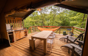 Safari tent with wooden deck, hammock, outdoor kitchen, dining table, chairs, and forest view.