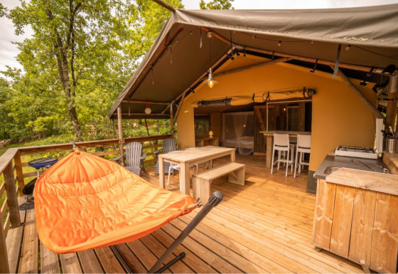Terraza exterior de tienda safari con muebles de madera, silla hamaca naranja y vista al bosque.