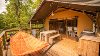 Terraza exterior de tienda safari con muebles de madera, silla hamaca naranja y vista al bosque.