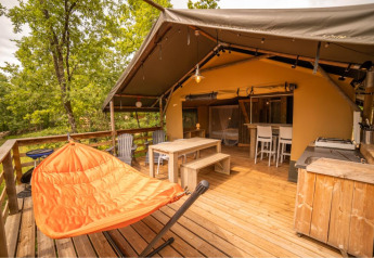 Outdoor deck of a safari tent with wooden furniture, orange hammock chair, and forest views.