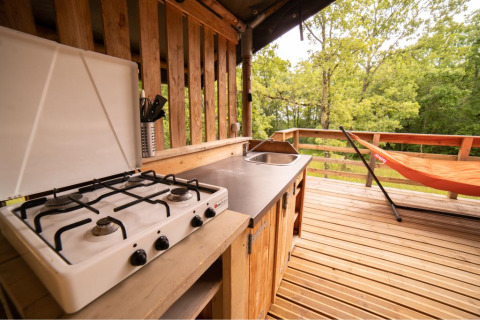 Buitenkeuken met gasvuur, spoelbak en oranje hangmat op houten terras naast een safaritent.