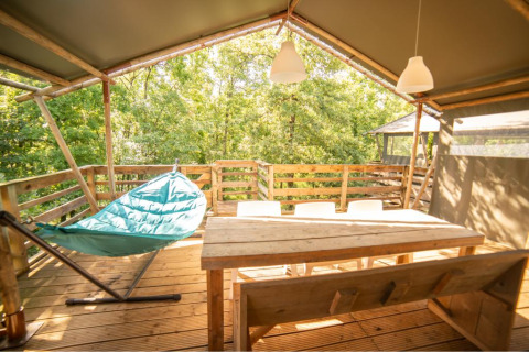 Terraza soleada de madera con hamaca, mesa y sillas en tienda safari Wood Lodge, Camping Les 3 Cantons, Francia