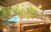 Terraza soleada de madera con hamaca, mesa y sillas en tienda safari Wood Lodge, Camping Les 3 Cantons, Francia