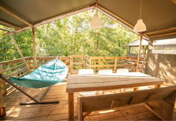 Terraza soleada de madera con hamaca, mesa y sillas en tienda safari Wood Lodge, Camping Les 3 Cantons, Francia