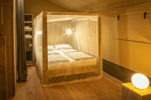 Camera da letto nella tenda safari Wood Lodge al Camping Les 3 Cantons in Francia, con zanzariera e pavimento in legno.