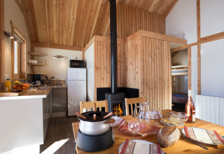 Intérieur de glamping chaleureux avec murs en bois, table dressée, poêle à bois et cuisine moderne au fond.