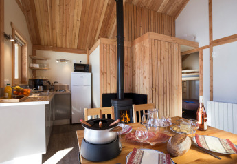 Sfeervolle glamping-accommodatie met houten interieur, gedekte tafel, haardvuur en moderne keuken.