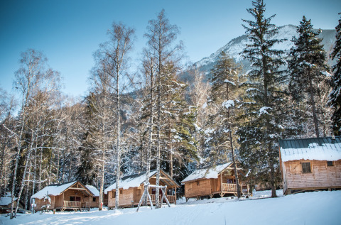 Snow-covered wooden cabins at Les Chalets Huttopia de Bozel, nestled among trees and mountains in Auvergne-Rhône-Alpes.