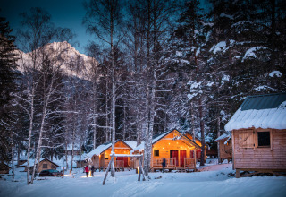 Beleuchtete Holzchalets im verschneiten Wald am Abend, Les Chalets Huttopia de Bozel in Auvergne-Rhône-Alpes.