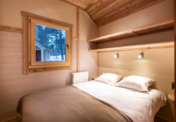 Cozy glamping room featuring a double bed, wooden interior, shelves, and a window with a forest view.