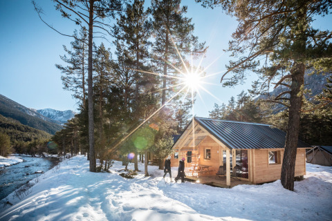 Cabaña de madera junto a un río nevado con sol, montañas y árboles, foto tomada en una posada.