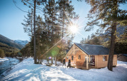 Houten chalet aan een besneeuwde rivier met zon, bomen en bergen, genomen bij een lodge.