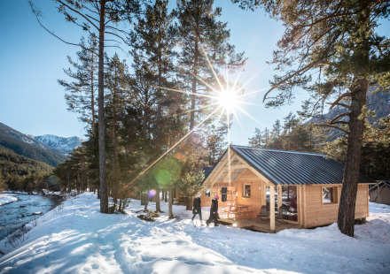 Houten blokhut aan een besneeuwde rivier met zonlicht, bergen en bomen, vastgelegd bij een lodge.