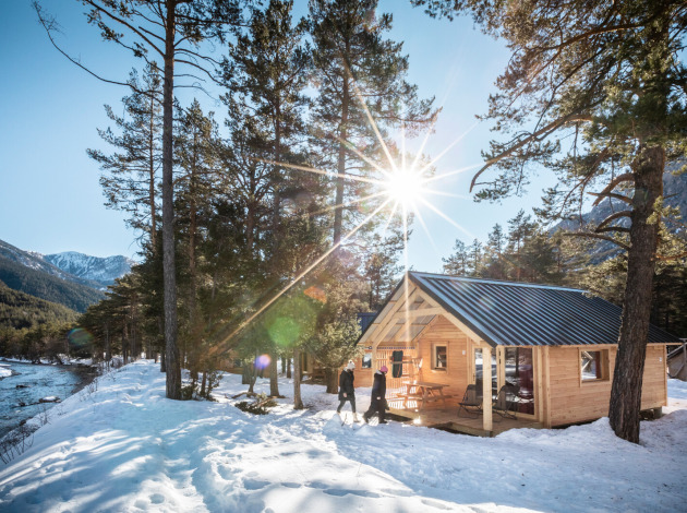 Houten blokhut aan een besneeuwde rivier met zonlicht, bergen en bomen, vastgelegd bij een lodge.