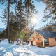 Træhytte ved en flod om vinteren med solskin, bjerge og sneklædte træer, set ved en lodge.