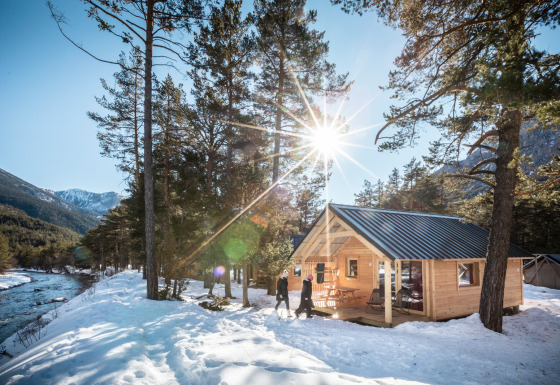Wooden cabin by a snowy river with sunlight shining through trees and mountains, taken at a lodge.