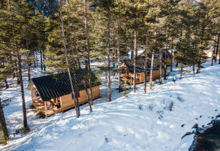 Twee houten chalets in een besneeuwd bos aan een rivier in Huttopia la Clarée, Provence-Alpes-Côte d’Azur.