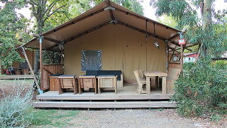 Tenda safari con arredi in legno sulla veranda, circondata da alberi, ideale per rilassarsi all'aperto.