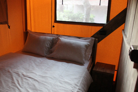 Schlafzimmerinterieur in einem Safarizelt mit Doppelbett, Kissen und gemütlicher Bettwäsche unter einem Fenster.