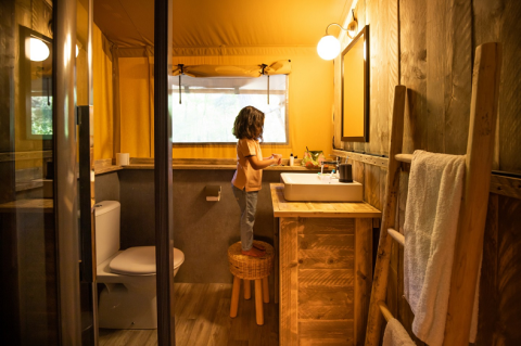Un niño se sube a un taburete en el baño acogedor de un alojamiento glamping de madera y luz cálida.