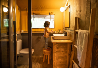Un niño se sube a un taburete en el baño acogedor de un alojamiento glamping de madera y luz cálida.