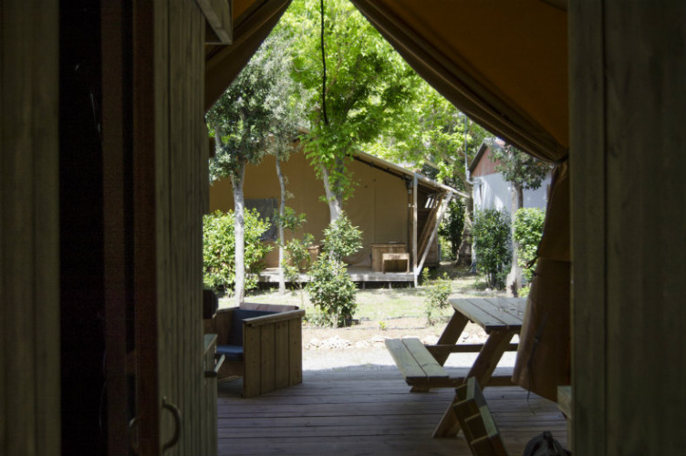 View from inside a safari tent with wooden benches, greenery, and a patio at Campo dei Fiori, Italy.