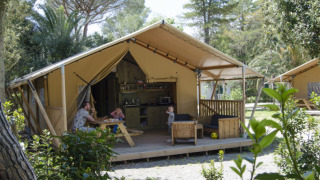 Family relaxing outside a safari tent with a kitchen and sitting area, surrounded by lush greenery.