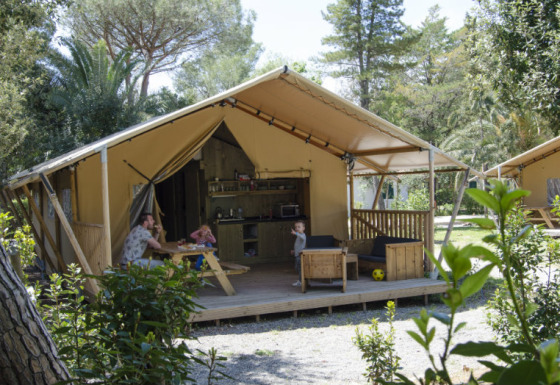 Famille se détend devant une tente safari avec cuisine et salon, entourée de verdure abondante.