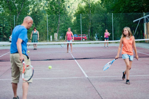 Familias juegan al tenis en una pista al aire libre rodeada de árboles en un parque vacacional en Francia.