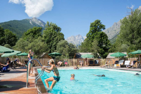 Outdoor pool at Huttopia Vallouise with people relaxing, green umbrellas, and mountain views in the background.