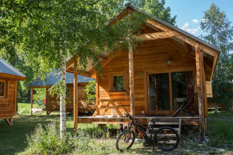 Cabaña de madera con bicicleta delante, rodeada de árboles en Huttopia Vallouise, Provenza-Alpes-Costa Azul, Francia.