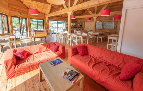 Sala luminosa en Huttopia Vallouise con sofás rojos, interior de madera y amplias ventanas con vista al exterior.