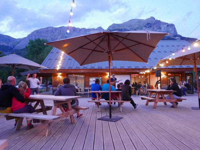 Außenterrasse mit Gästen, Lichterketten, Sonnenschirmen und Bergblick im Huttopia Vallouise, Frankreich.