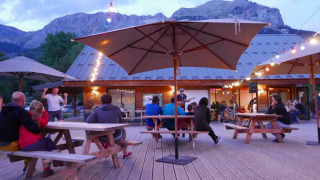 Zona de descanso al aire libre con huéspedes, luces, sombrillas y vista a las montañas en Huttopia Vallouise.