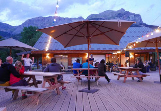 Outdoor seating area with guests, string lights, umbrellas, and mountain views at Huttopia Vallouise, France.