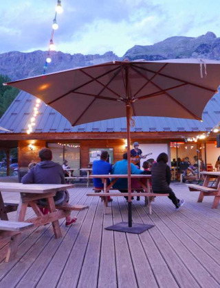 Zona de descanso al aire libre con huéspedes, luces, sombrillas y vista a las montañas en Huttopia Vallouise.