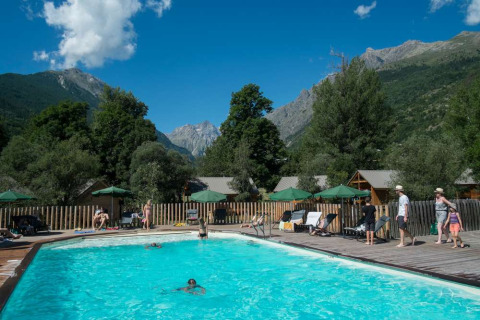 Piscina al aire libre con huéspedes, montañas y cabañas de fondo en Huttopia Vallouise, Francia.