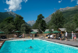Outdoor swimming pool with guests, mountains, and cabins in the background at Huttopia Vallouise, France.