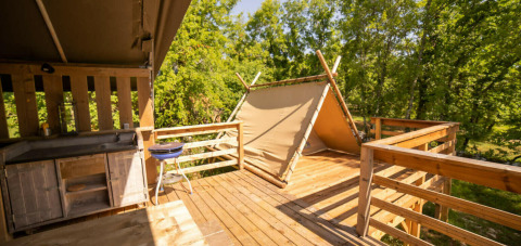 Terraza de madera con cocina y pequeña tienda en Safari Lodge, Camping Les 3 Cantons, Francia, rodeada de árboles.